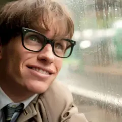 Junger Stephen Hawking mit Brille blickt lächelnd aus einem Fenster, das von Regentropfen bedeckt ist.