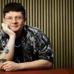Fritz Espenlaub, mit Brille und Tattoos, lehnt nachdenklich am Tisch vor einer vertikalen Wand.