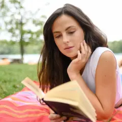 Junge Frau liest auf einer Decke im Gras ein Buch. Sie ist vertieft in die Geschichte, die sie gerade liest, und genießt die Ruhe der Natur. Ein perfekter Tag für gute Romane.