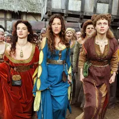 Drei Frauen in historischen Gewändern, die an eine "die wanderhure reihenfolge" erinnern, gehen durch eine mittelalterliche Gasse.
