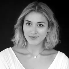 Porträt von Katharina Fuchs, die in die Kamera blickt. Sie trägt ein weißes Oberteil und eine zarte Halskette.