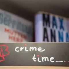 Ein Schild mit der Aufschrift "crime time" und einem roten Logo. Im Hintergrund unscharfe Bücher, die auf gemütliche Krimi-Lektüre hindeuten.