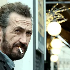 Rocco Schiavone, mit zerknittertem Gesicht und Zigarette im Mund, blickt nachdenklich in die Ferne.