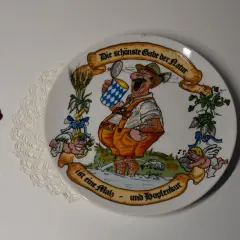 Ein Teller mit einem fröhlichen Mann im Lederhosen, der ein Bier hält. Die Inschrift lautet: "Die schönste Gabe der Natur ist eine Malz- und Hopfenkur." Judith Merkle Riley hat dieses Motiv entworfen.