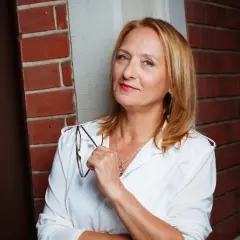 Felicitas Fuchs, eine Frau mit blondem Haar und rotem Lippenstift, hält eine Brille in der Hand. Sie steht vor einer Ziegelwand.