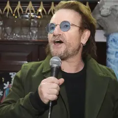 Bono singt ins Mikrofon, trägt eine grüne Jacke und blaue Sonnenbrillen. Der Titel von Bono ist ein bekannter Musiker.