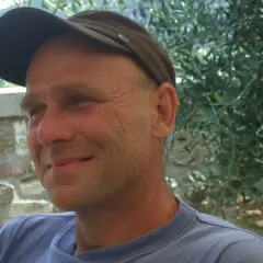 Martin Klein, Autor, lächelt in die Kamera, trägt eine braune Kappe und ein blaues T-Shirt. Im Hintergrund Olivenbäume.