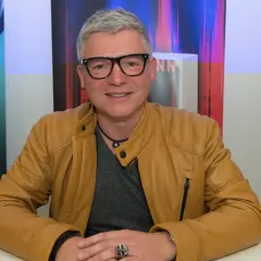 Andreas Winkelmann sitzt lächelnd in einer braunen Lederjacke. Hinter ihm sind bunte Banner zu sehen, die auf seine Bücher und Lesungen hinweisen.