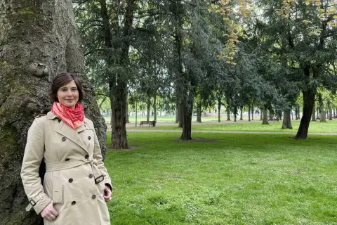 Kristina Hartenbach lehnt an einem Baum im Park. Sie trägt einen beigen Trenchcoat und einen roten Schal.