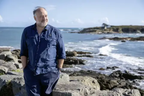 Ein Mann in blauer Kleidung steht am Meer. Die bannalec reihenfolge der Wellen spiegelt sich in seiner entspannten Haltung wider.