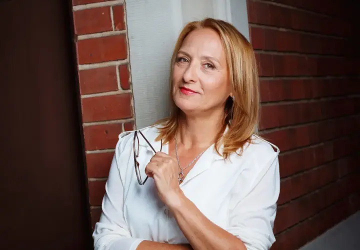 Felicitas Fuchs, eine Frau mit blondem Haar und rotem Lippenstift, hält eine Brille in der Hand. Sie steht vor einer Ziegelwand.