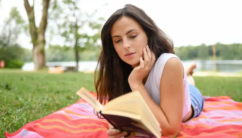 Junge Frau liest auf einer Decke im Gras ein Buch. Sie ist vertieft in die Geschichte, die sie gerade liest, und genießt die Ruhe der Natur. Ein perfekter Tag für gute Romane.