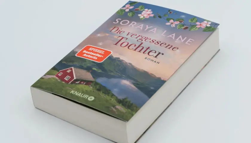 Ein Buchcover mit dem Titel "Die vergessene Tochter" von Soraya Lane. Es zeigt eine rote Hütte am See vor einer Bergkulisse.