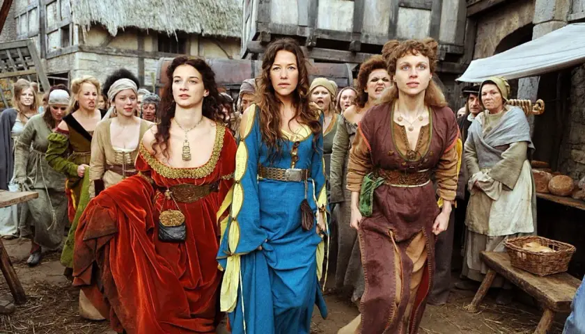Drei Frauen in historischen Gewändern, die an eine "die wanderhure reihenfolge" erinnern, gehen durch eine mittelalterliche Gasse.