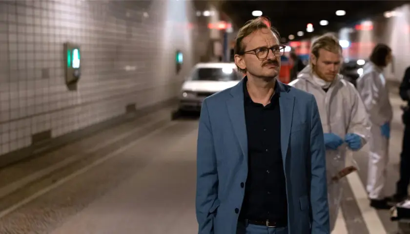 Ein Mann im blauen Anzug, vielleicht ein Ermittler wie Danowski, steht im Tunnel. Im Hintergrund arbeiten Personen in Schutzanzügen.