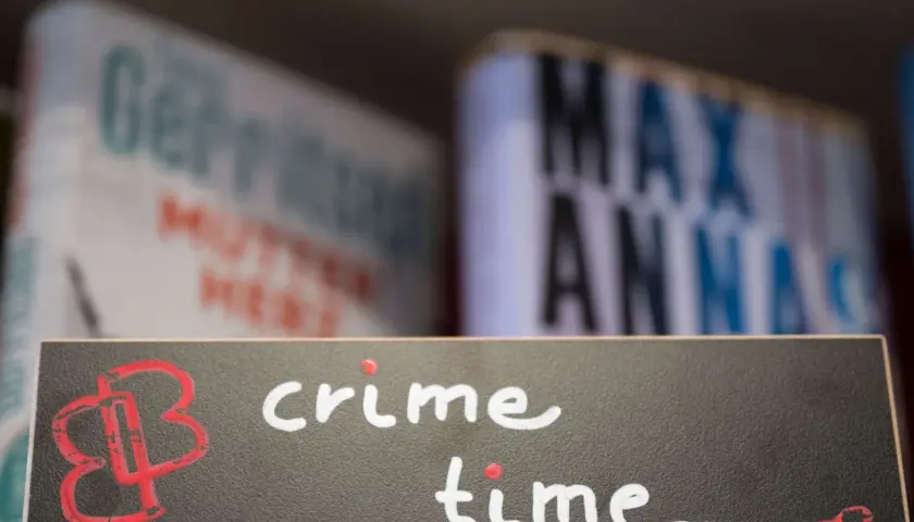 Ein Schild mit der Aufschrift "crime time" und einem roten Logo. Im Hintergrund unscharfe Bücher, die auf gemütliche Krimi-Lektüre hindeuten.