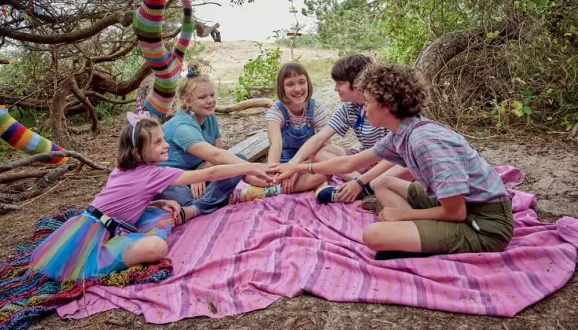 Fünf Kinder legen ihre Hände in der Mitte zusammen, bereit für ein Abenteuer. Die bunte Strickkunst am Baum deutet auf die "lotta leben reihenfolge" hin, die sie gemeinsam entdecken werden.
