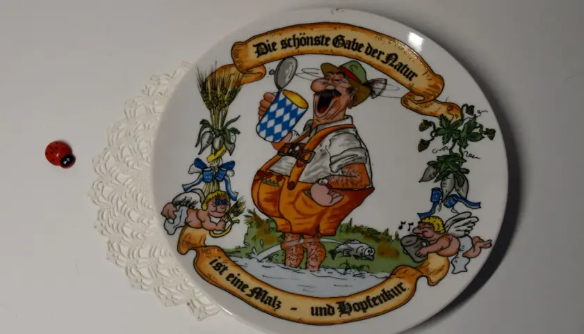 Ein Teller mit einem fröhlichen Mann im Lederhosen, der ein Bier hält. Die Inschrift lautet: "Die schönste Gabe der Natur ist eine Malz- und Hopfenkur." Judith Merkle Riley hat dieses Motiv entworfen.