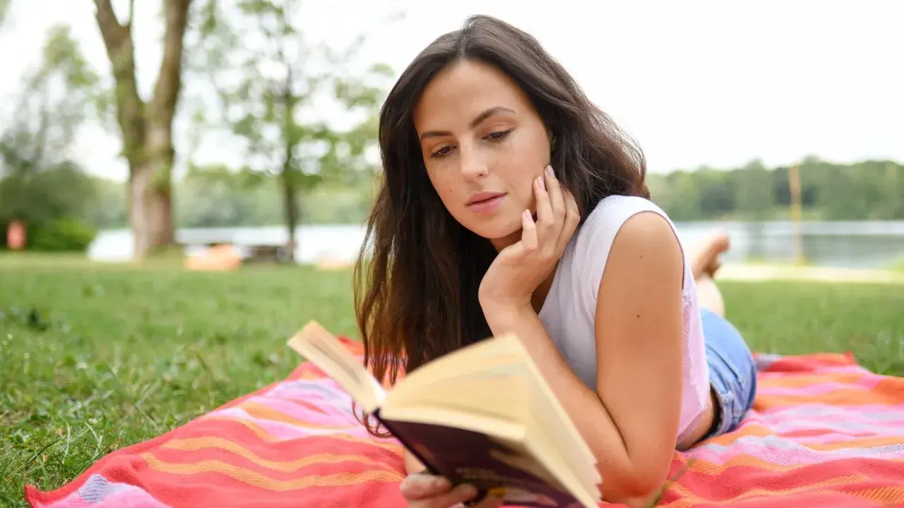 Junge Frau liest auf einer Decke im Gras ein Buch. Sie ist vertieft in die Geschichte, die sie gerade liest, und genießt die Ruhe der Natur. Ein perfekter Tag für gute Romane.