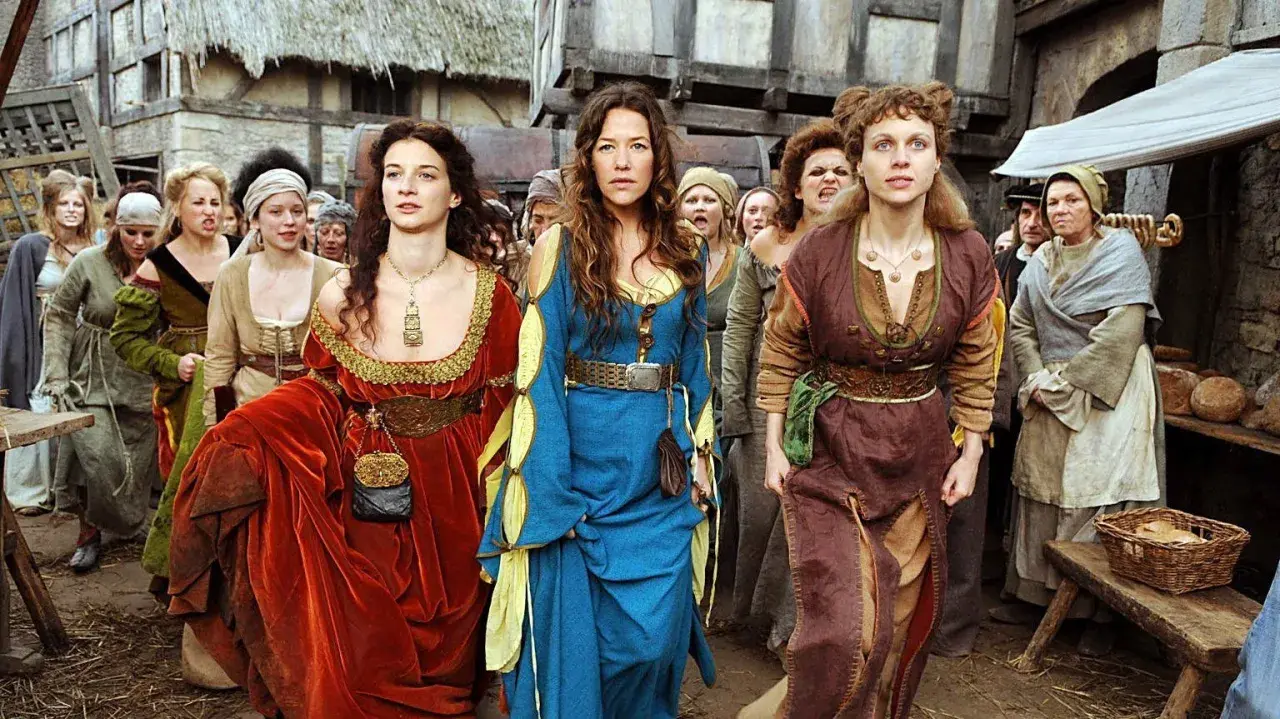Drei Frauen in historischen Gewändern, die an eine "die wanderhure reihenfolge" erinnern, gehen durch eine mittelalterliche Gasse.