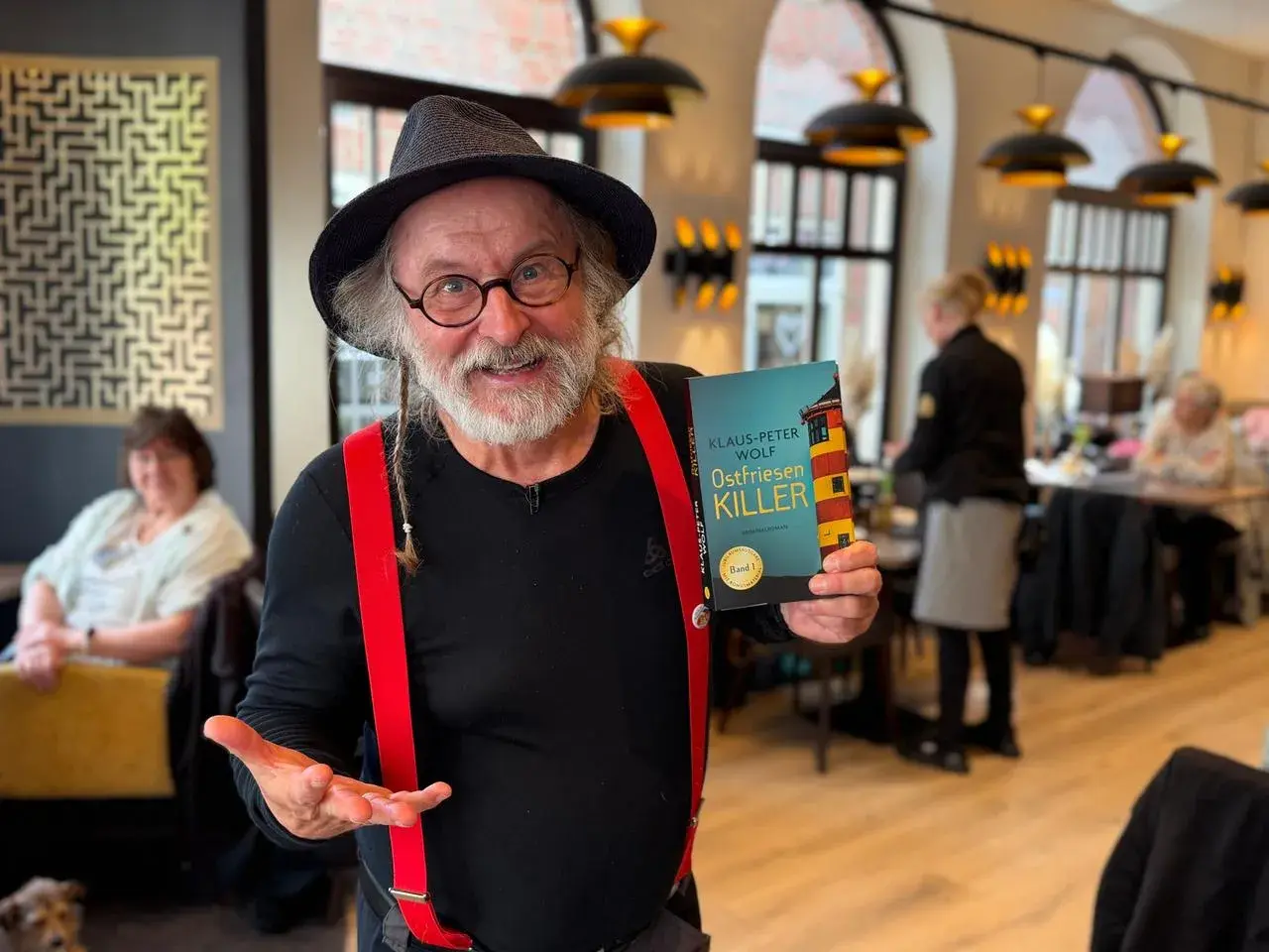 Klaus-Peter Wolf präsentiert "Ostfriesenkiller", Band 1 seiner Krimireihe. Ein Mann mit Hut und roten Hosenträgern hält das Buch hoch.