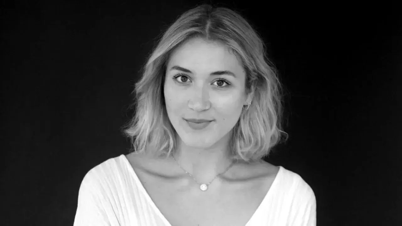 Porträt von Katharina Fuchs, die in die Kamera blickt. Sie trägt ein weißes Oberteil und eine zarte Halskette.