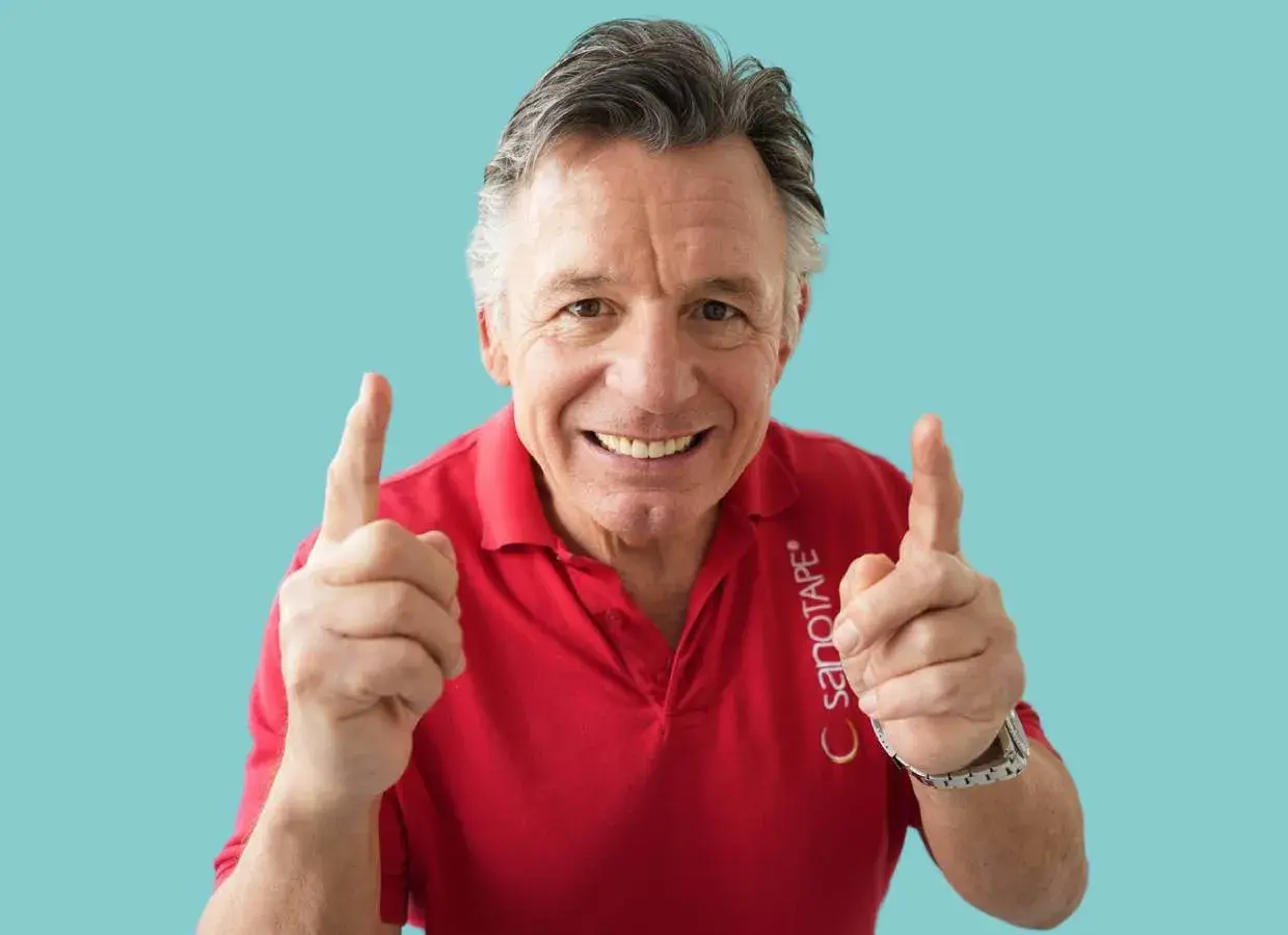 Steffen Barth zeigt mit beiden Zeigefingern nach oben und lächelt. Er trägt ein rotes Poloshirt mit dem Aufdruck "SANOTAPE".