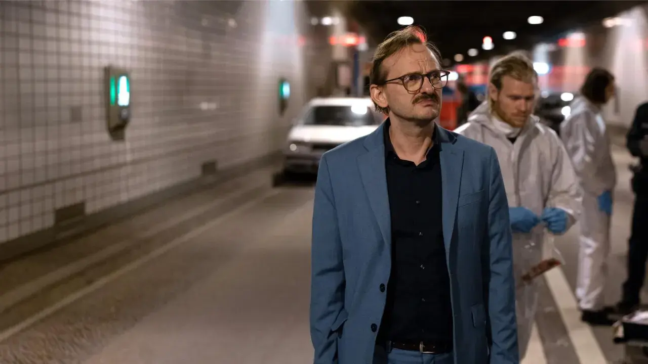 Ein Mann im blauen Anzug, vielleicht ein Ermittler wie Danowski, steht im Tunnel. Im Hintergrund arbeiten Personen in Schutzanzügen.