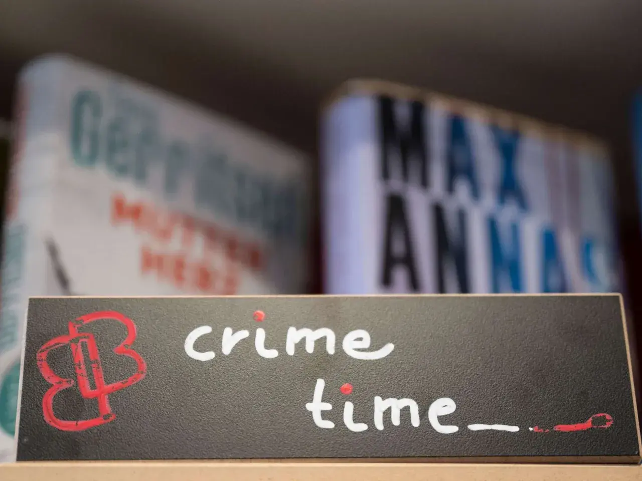Ein Schild mit der Aufschrift "crime time" und einem roten Logo. Im Hintergrund unscharfe Bücher, die auf gemütliche Krimi-Lektüre hindeuten.