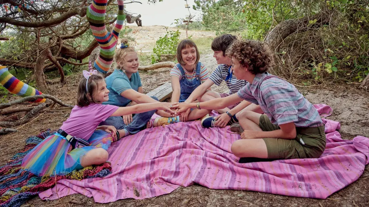 Fünf Kinder legen ihre Hände in der Mitte zusammen, bereit für ein Abenteuer. Die bunte Strickkunst am Baum deutet auf die "lotta leben reihenfolge" hin, die sie gemeinsam entdecken werden.