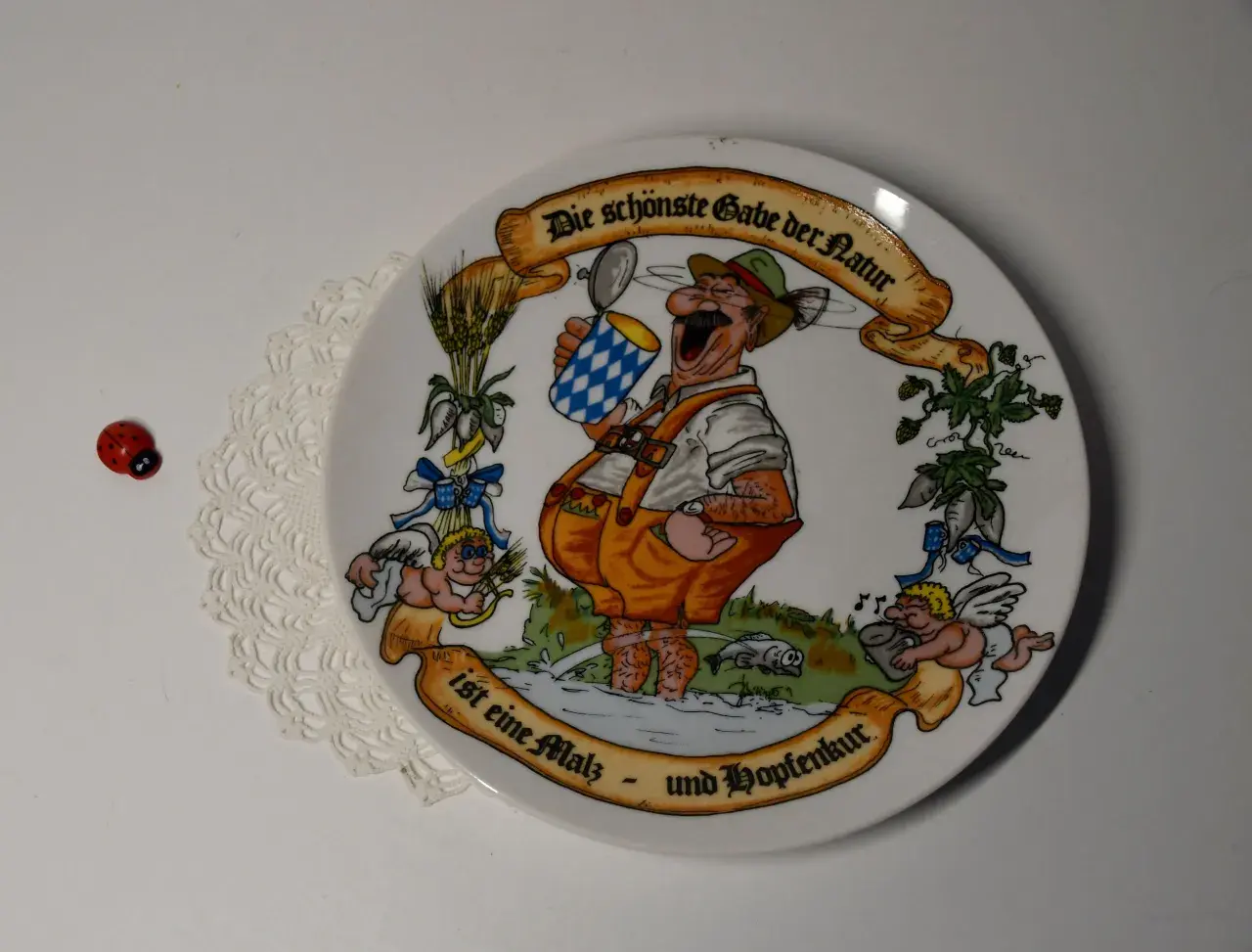 Ein Teller mit einem fröhlichen Mann im Lederhosen, der ein Bier hält. Die Inschrift lautet: "Die schönste Gabe der Natur ist eine Malz- und Hopfenkur." Judith Merkle Riley hat dieses Motiv entworfen.