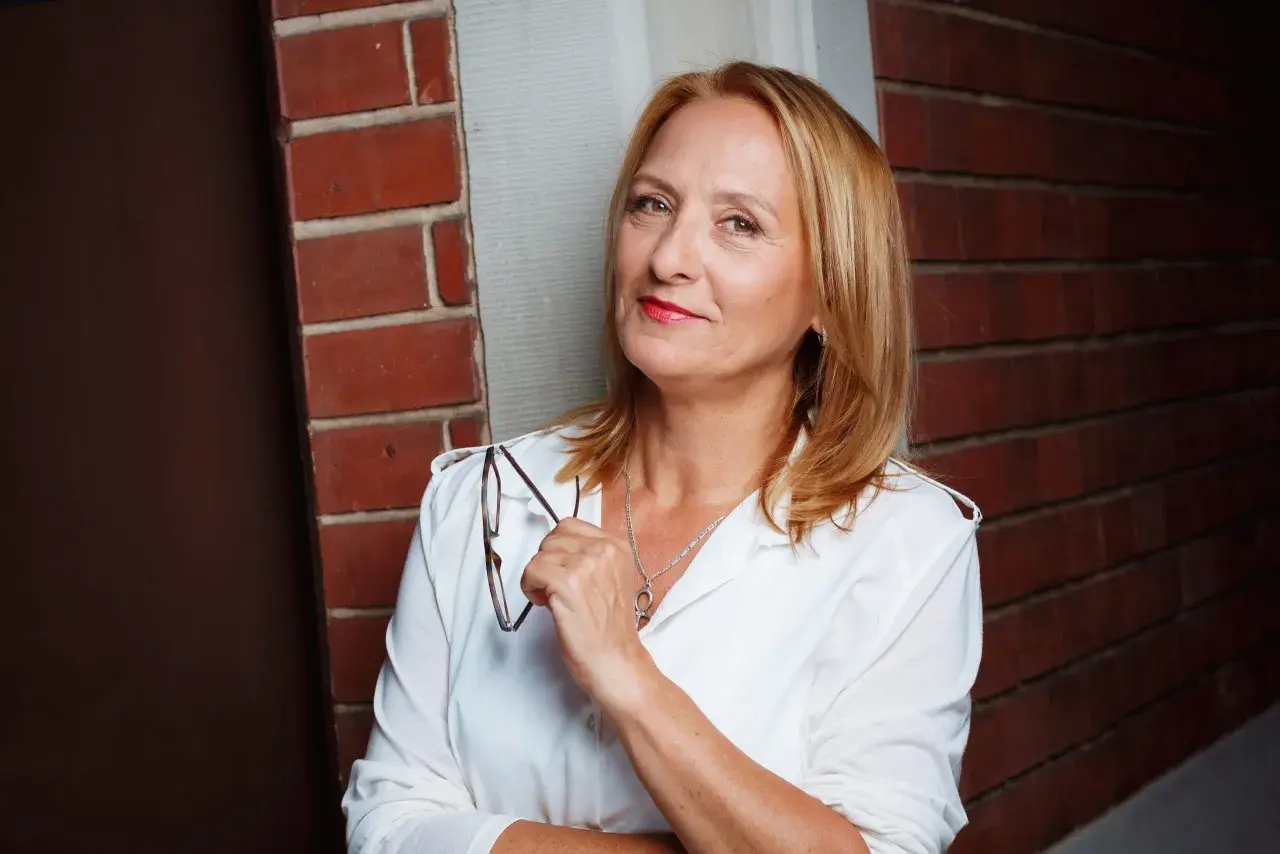 Felicitas Fuchs, eine Frau mit blondem Haar und rotem Lippenstift, hält eine Brille in der Hand. Sie steht vor einer Ziegelwand.