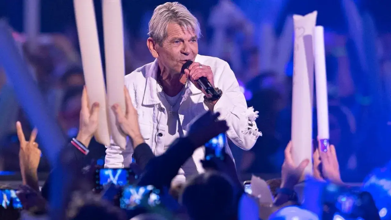 Ein Sänger singt auf der Bühne, umgeben von Fans mit Lichtern. Er scheint zu sagen: "Wie lieb ich dich hab", und die Menge jubelt.