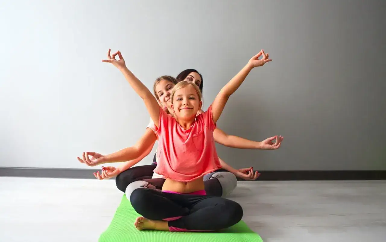 Drei Mädchen üben Yoga. Sie sitzen im Schneidersitz auf einer grünen Matte und machen eine Handgeste. Yoga-geschichten für kinder zum mitmachen.