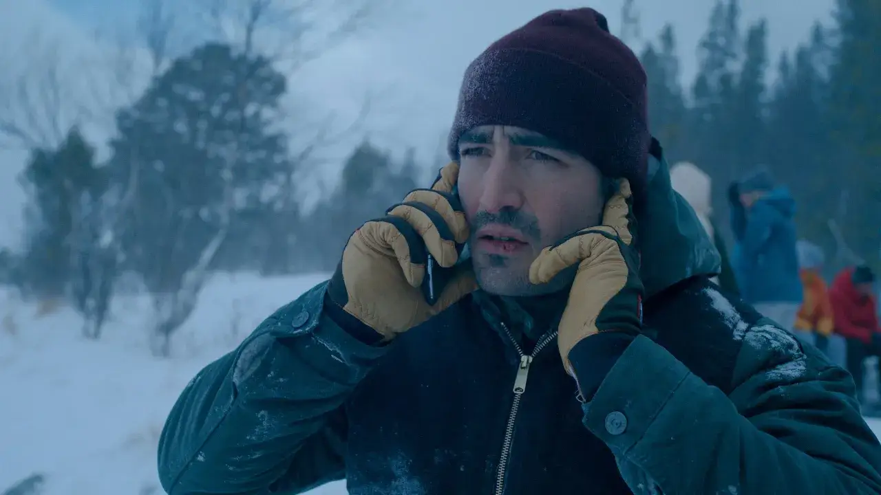 Mann mit blutiger Lippe telefoniert im Schnee, typisch für nordic noir. Im Hintergrund verschwommene Gestalten und Bäume.
