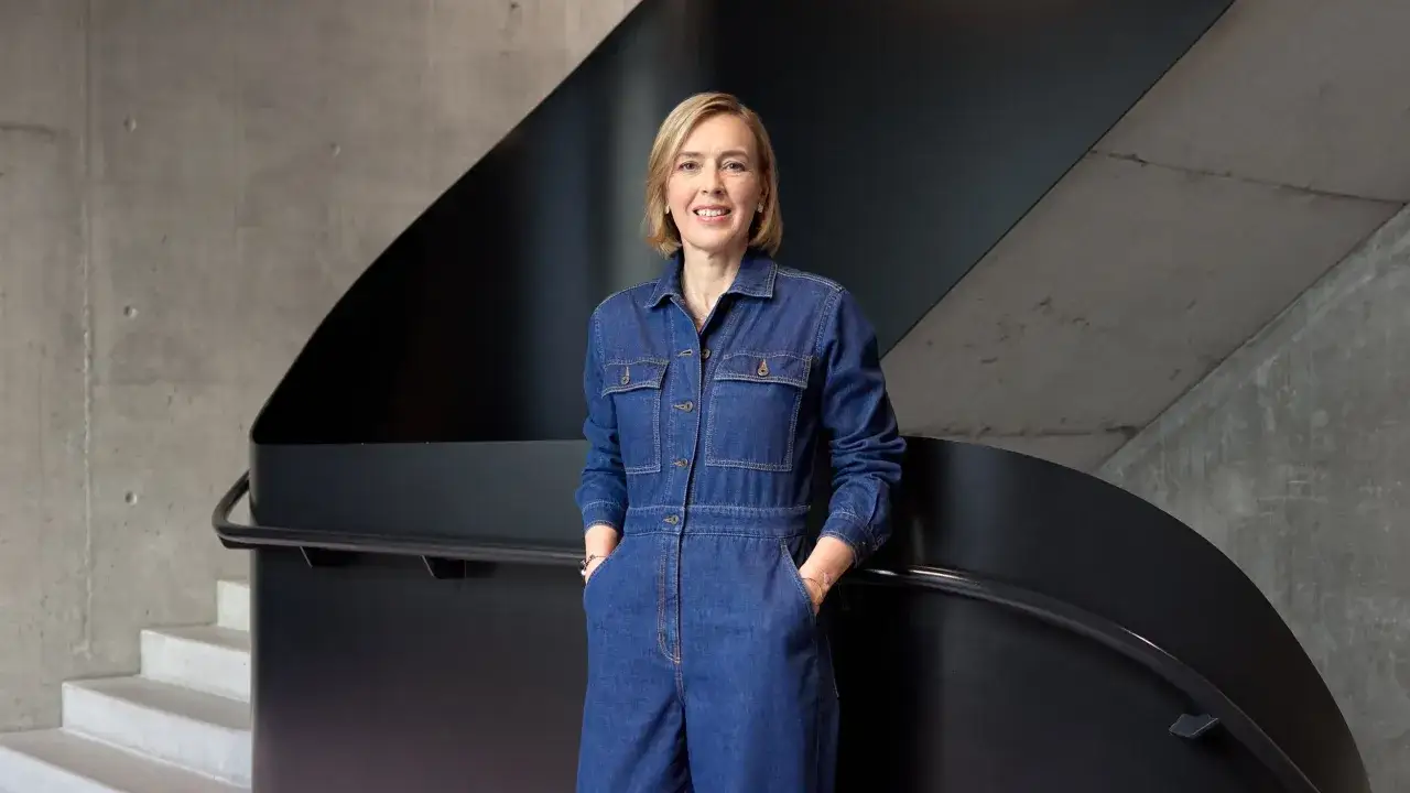 Christiane von Hardenberg lächelt in einem Jeans-Overall, die Hände in den Taschen, vor einer modernen Treppe aus Beton und schwarzem Metall.