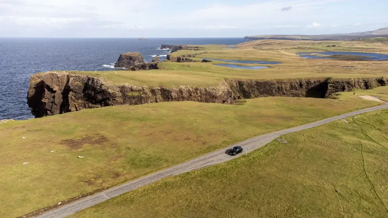 Ein schwarzes Auto fährt auf einer einsamen Straße entlang der dramatischen Klippen der Shetlandinseln. Ein perfekter Schauplatz für einen Shetland Krimi.